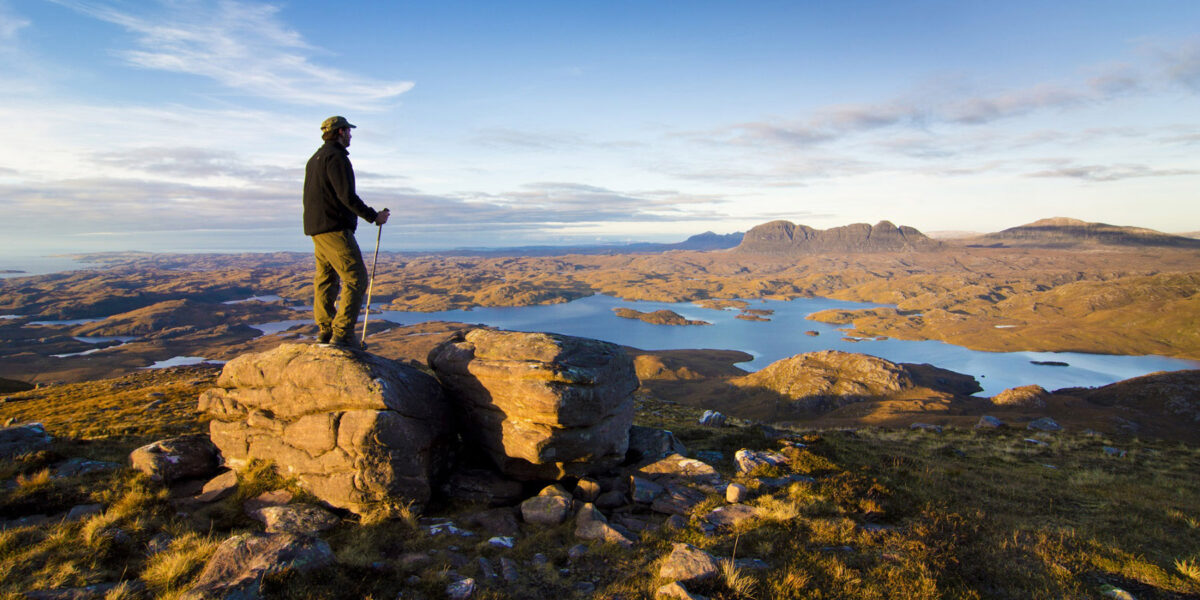 Ein Mensch steht mit Wanderstöcken auf einem Berg in Schottland und überblickt eine steinige, bergige Landschaft in welcher auch ein See abgebildet ist.