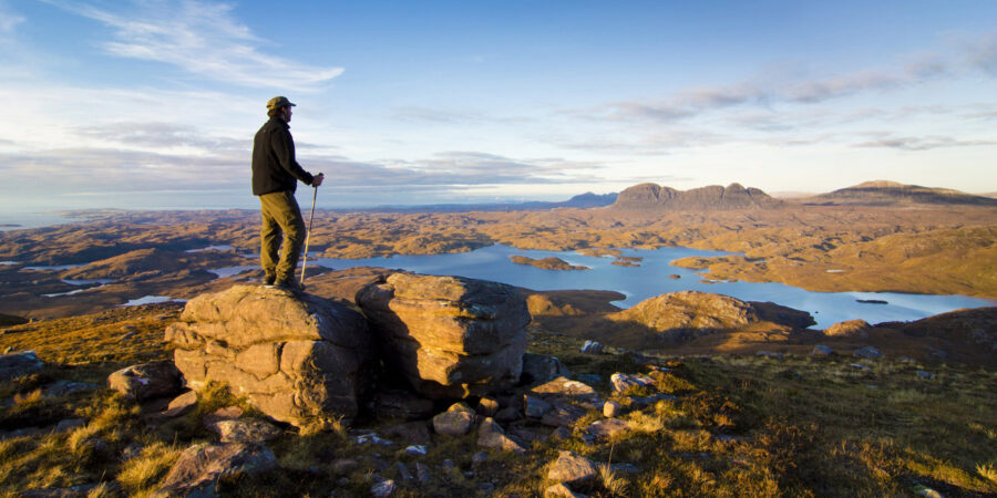 Ein Mensch steht mit Wanderstöcken auf einem Berg in Schottland und überblickt eine steinige, bergige Landschaft in welcher auch ein See abgebildet ist.