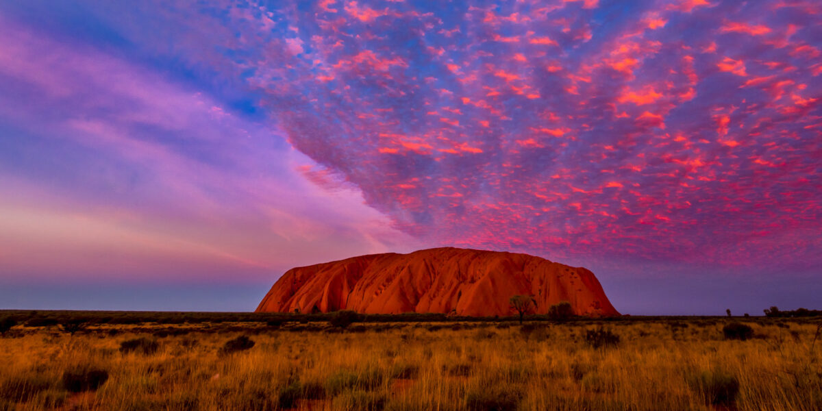 Abgebildet ist der Ayer`s Rock in Australien in einem violett-pinken Sonnenuntergangszenario.