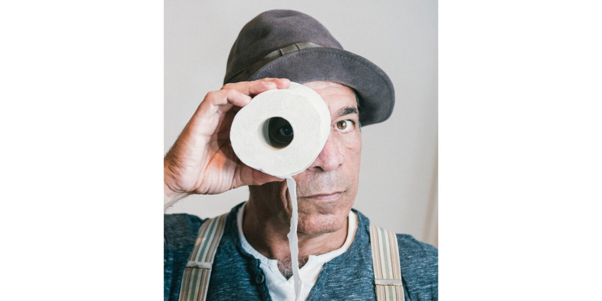 Peter Shub in einem Portraitfoto. Er trägt seine Mütze und schaut mit einem Auge durch eine Klorolle in die Kamera.