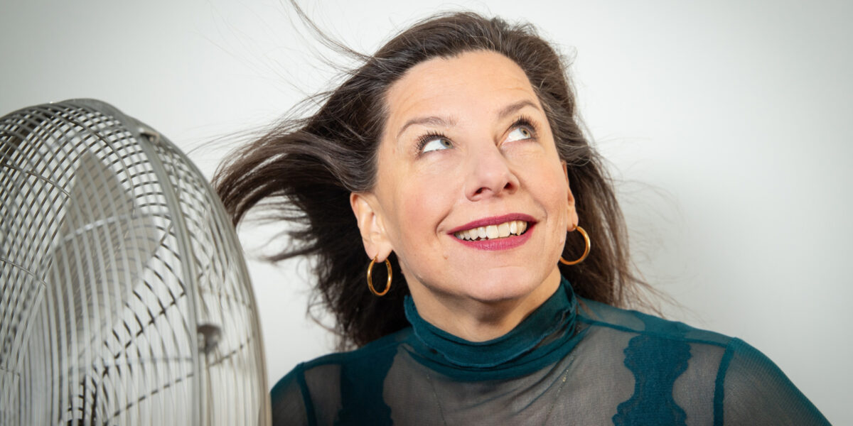 Eine Frau schaut in die Ecke oben rechts. Links steht ein Ventilator, der ihre Haare zum Wehen bringt.