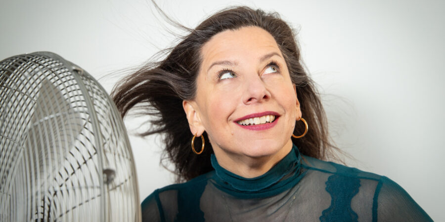 Eine Frau schaut in die Ecke oben rechts. Links steht ein Ventilator, der ihre Haare zum Wehen bringt.