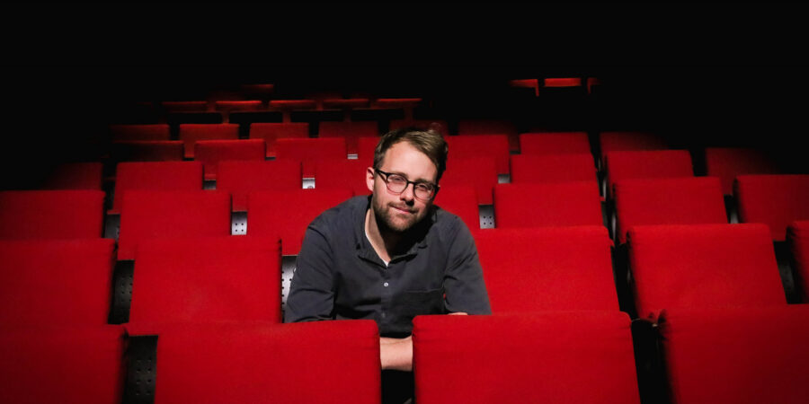 Lars Ruppel sitzt inmitten einer Reihenbestuhlung von roten Stühlen, wie man sie aus einem Kino kennt. Er schaut in die Kamera.